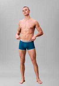 Turkusowe bokserki z szarą gumką Calvin Klein, dopasowany fason, miękka tekstura i długość sięgająca powyżej kolan, noszone przez męskiego modela bez koszulki.