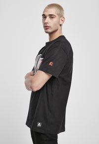 Černé bavlněné tričko s grafickým potiskem na přední straně, krátkými rukávy a červeným logem na levém rukávu.