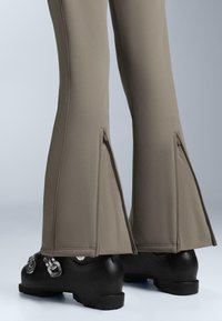 Pantaloni a zampa d'elefante in tessuto grigio chiaro con cerniere laterali, abbinati a stivali neri con fibbie metalliche e suola texturizzata.