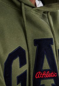 Sweatshirt vert olive avec capuche, présentant un grand logo "GAP" bleu foncé et le texte "Athletic" rouge, fabriqué en tissu doux avec une finition texturée.