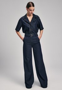 Camicia in denim scuro con colletto e chiusura a bottoni, abbinata a pantaloni a vita alta e gamba larga. Presenta cuciture a vista e una cintura.