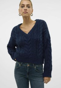 Navy blue knitted sweater with a deep V-neck, featuring a textured cable pattern and ribbed hem. Paired with dark jeans.