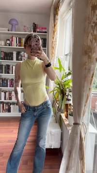 Gul toppløs bodysuit med høy hals, kombinert med lys blå flagrende jeans. Bakgrunnen har hyller med bøker og planter.