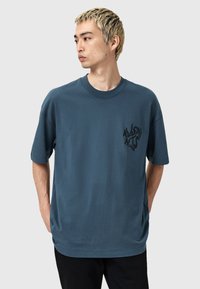 Camiseta de algodón azul con mangas cortas, cuello redondo y un logo gráfico negro en el lado izquierdo del pecho. Ajuste casual y relajado.