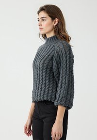 Maglione grigio lavorato a maglia con collo alto, caratterizzato da un motivo a treccia testurizzato e maniche a palloncino, abbinato a pantaloni neri. Nessun abbellimento visibile.