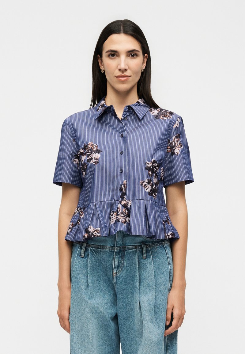 Femme aux longs cheveux foncés portant un chemisier rayé bleu à manches courtes avec broderies florales et ourlet volanté, associé à un jean en denim taille haute.
