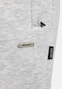 Szare dresy wykonane z teksturowanej tkaniny, z małą srebrną metką "SMILODOX" oraz czarną etykietą logo po boku.
