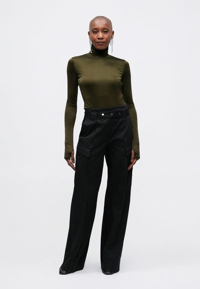 Top verde măslin cu guler înalt și mâneci lungi, asociat cu pantaloni negri cu talie înaltă, evazați, care au o talie cu curea și buzunare laterale.