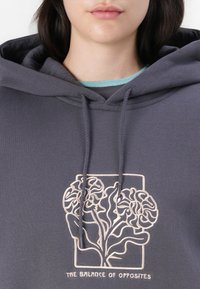 Grå hettegenser med frontlomme, utstyrt med en lys fersken brodert grafikk av et tre med svingende blader og teksten "BALANSEN MELLOM MOTSETNINGER."
