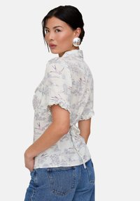 Lightweight, floral-patterned blouse with short ruffled sleeves, a tied back, and a collared neckline, paired with high-waisted jeans.