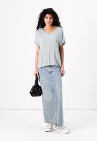 Lys grå løs bluse med V-hals, parret med en lang denimskørt. Modellen holder en sort blomster taske og har hvide sneakers på.