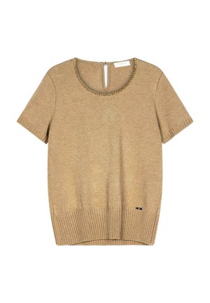 Jeune femme à la peau claire et aux longs cheveux bruns porte un haut en maille beige à manches courtes avec un détail chaîne au niveau du décolleté et un pantalon noir.