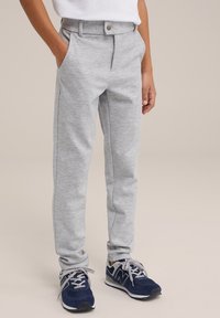 Dítě v světle šedých kalhotách, s rukama v kapsách a tmavě modrých teniskách, stojící na neutrálním pozadí.