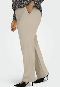 Bež hlače ravnog kroja s bočnim džepovima, izrađene od glatke tkanine. Nošene s uzorkovanom bluzom i crnim cipelama na visoku petu.