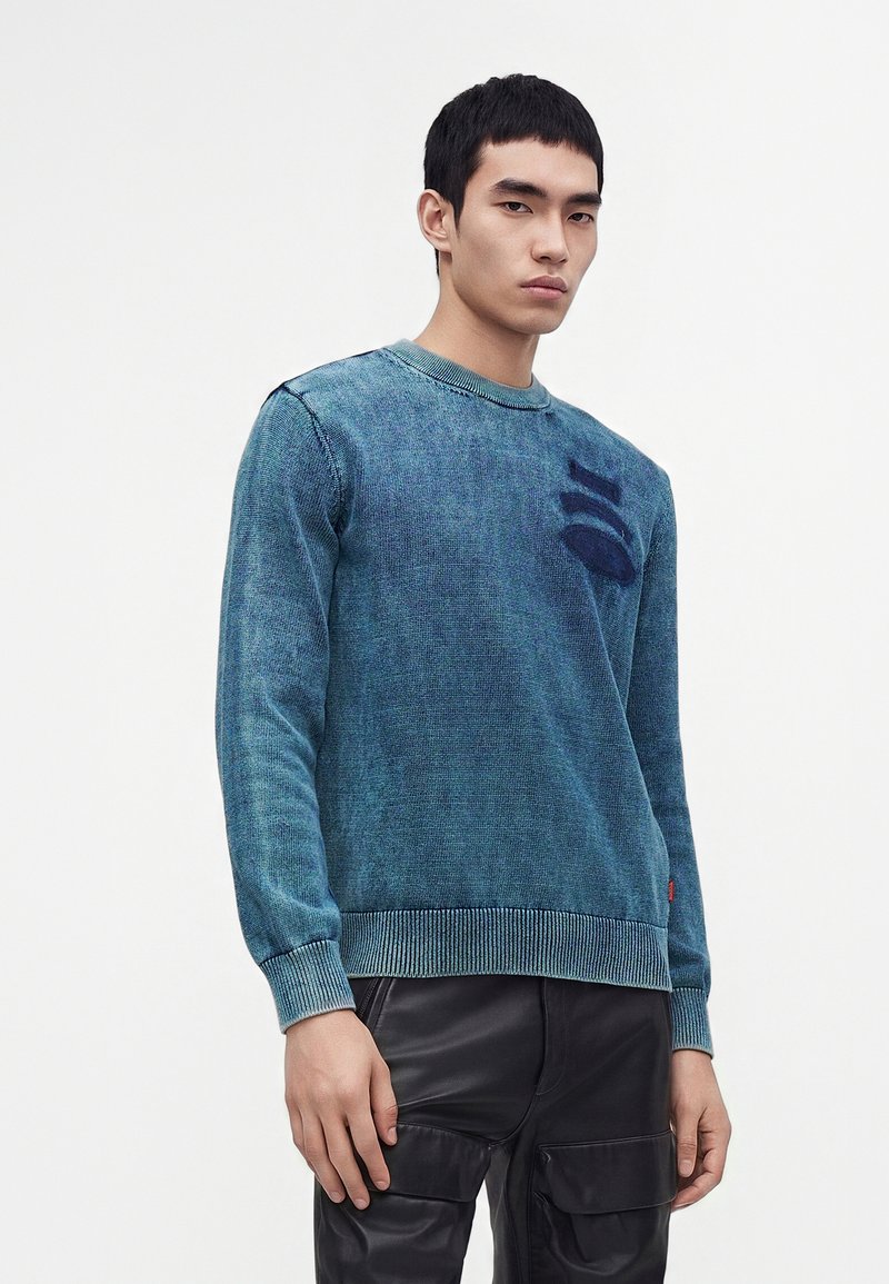 Jeune homme aux cheveux noirs courts, portant un pull en maille bleu-vert et un pantalon en cuir noir, debout devant un fond clair uni.