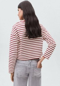 Kvinna med långt mörkt hår som bär en vit och röd randig långärmad tröja och ljusgrå jeans, stående med ena handen i bakfickan.
