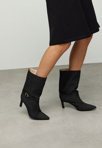 Bottes de cheville en tissu noir avec bouts pointus, talons stilettos, et une sangle noire ornée d'une boucle dorée à mi-mollet.