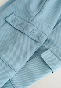 Helleblaue Textilie mit einer gerippten Struktur und einem rechteckigen Patch, auf dem das eingestanzte Wort "BAKER" in einer passenden Farbe zu sehen ist.