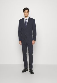 Marineblaues Sakko mit zwei Knöpfen, Reverskragen und einer einzelnen Brusttasche. Passende Hose. Weißes Hemd und marineblauer Krawatte. Schwarze Oxfords.