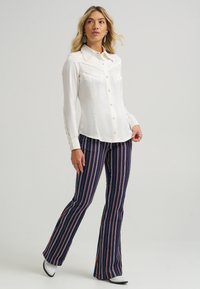 Hvit bluse med knapper, krage og dekorative sting, kombinert med marineblå, stripete vide bukser med oransje detaljer.