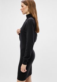 Vestido de malha preto com um decote alto, mangas longas e silhueta ajustada, apresentando uma textura suave e punhos canelados.