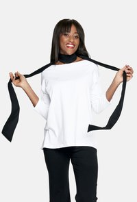 Biała bluzka z długim rękawem i dekoltem łódkowym, zestawiona z czarnymi spodniami. Czarny szal z tkaniny luźno opadający na szyi i rękach.