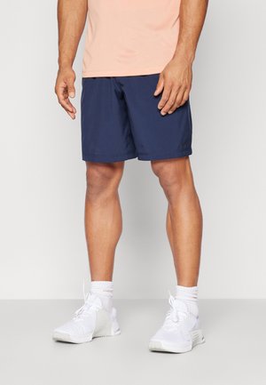 Un mannequin porte un short de sport bleu marine, un haut couleur pêche, des chaussettes blanches et des chaussures de course blanches sur un fond de studio uni.