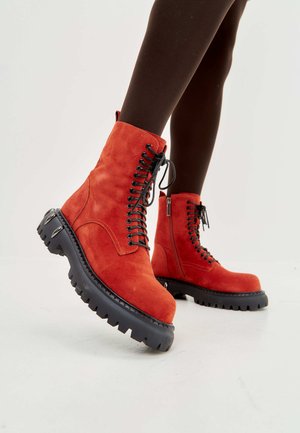 Bottines en suede rouge avec des lacets noirs, semelle épaisse en caoutchouc noir et accents en métal argenté. Fermeture à glissière sur le côté pour un enfilage facile.