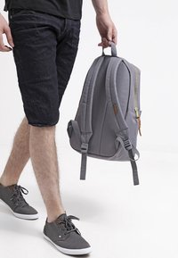 Mochila gris con doble cinta para los hombros, cierre de cremallera y acentos de cuero. Hecha de tela, con forma redondeada y un diseño minimalista.