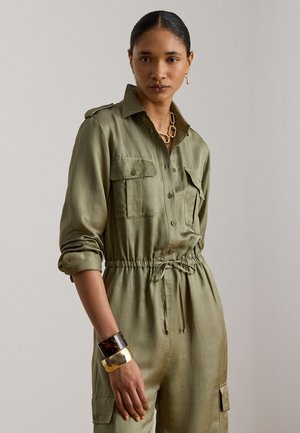Kvinde iført olivengrøn jumpsuit med knapper, opsmøgede ærmer, to brystlommer og snøre i taljen, accessoriseret med chunky armbånd og halskæde.