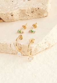 Paar goldene Steckohrringe mit kleinen bunten Steinen und ein Paar goldene Creolen mit hängenden klaren Edelsteinen auf beigen Steinscheiben.