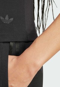 Musta urheilukangas esittelee tyylikkään muotoilun sivuraidoilla ja pienellä logolla. Kankaan pinta on sileä, ja se korostaa muotoon ommeltua siluettia.