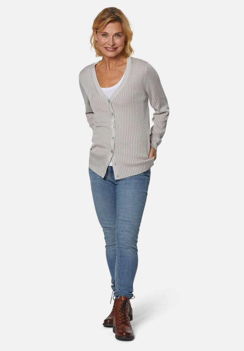 Lichtgrijze geribbelde cardigan met knopen, gedragen over een wit shirt. Gecombineerd met blauwe jeans en bruine veterschoenen. Eenvoudig, textuurontwerp.