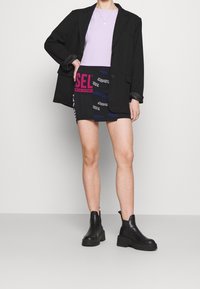 Zwarte blazer over een lila t-shirt, gecombineerd met een zwarte minirok met kleurrijke DIESEL-logo's. Zwarte leren enkellaarsjes.