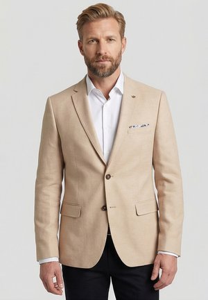 Man met blond haar en baard draagt een beige blazer over een wit overhemd met pochet, staand tegen een effen grijze achtergrond.