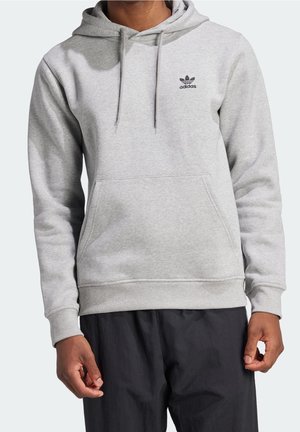 Mężczyzna noszący jasnoszarą bluzę Adidas z kapturem, z przednią kieszenią i czarnym logo, w połączeniu z luźnymi czarnymi spodniami na jednolitym tle.