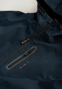 Veste bleu foncé avec capuche, avec deux fermetures éclair noires résistantes à l’eau et texte orange "001_089_247" près de chaque tirette.