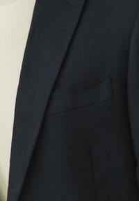 Mörkblå blazer i texturerat tyg, med en enfaldig bröstficka och skräddarsydda slag. Bärs över en ljusfärgad skjorta.