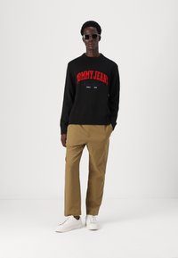 Svart genser med rød "TOMMY JEANS"-logo, beige bukser og hvite joggesko. Har rund hals og ribbestrikkede mansjetter. Minimalistiske solbriller.