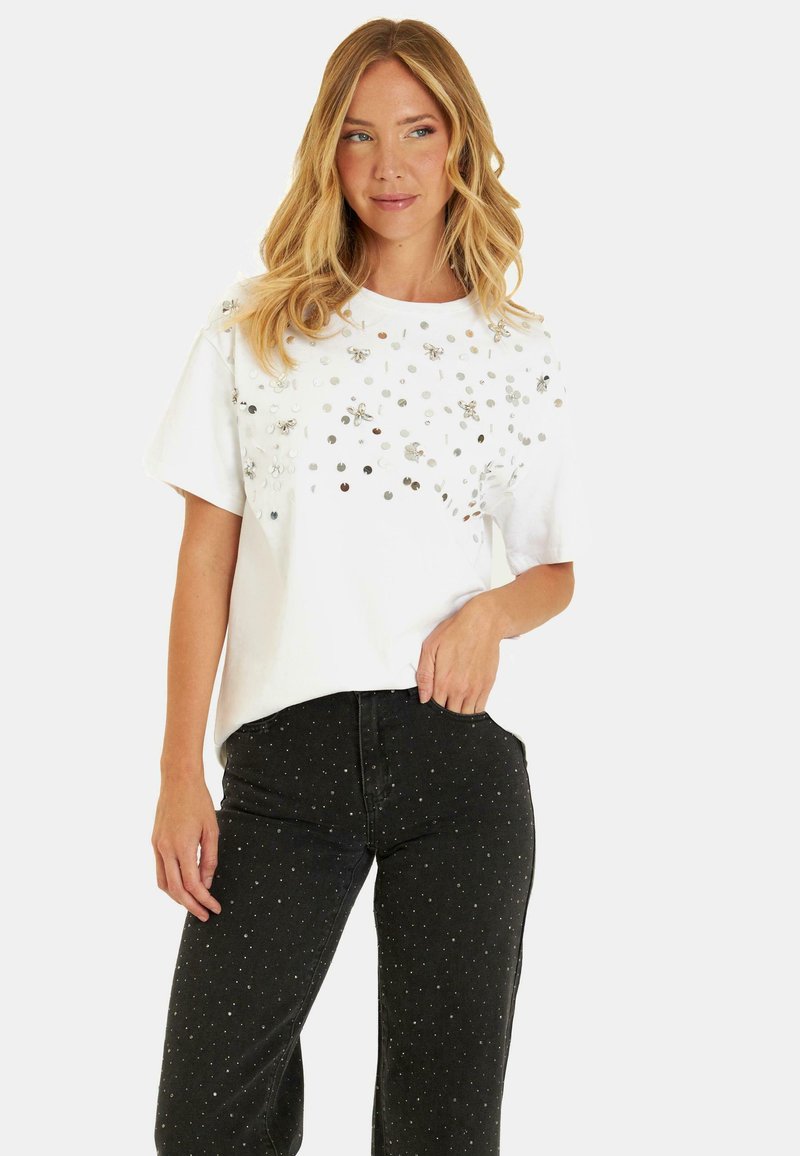 T-shirt blanc à manches courtes avec des embellissements métalliques sur le devant, associé à un jean noir avec des motifs de points argentés.