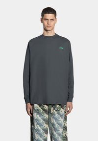 Szara koszulka z długim rękawem, z okrągłym dekoltem i zielonym logo "Prosto". W zestawieniu z spodniami w kamuflażu o postrzępionej fakturze.