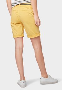 Pantalones cortos amarillos enrollados de algodón, con un bolsillo trasero y combinados con zapatillas blancas. Camiseta a rayas visible por encima de los pantalones cortos.