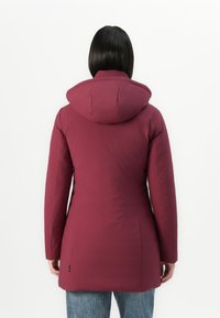 Vrouw met steil zwart haar in een lange bordeauxrode gewatteerde jas met capuchon, staand tegen een effen witte achtergrond, met de rug naar de camera gewend.
