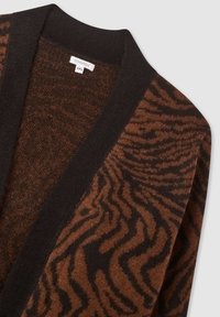 Brązowy kardigan we wzory z czarnymi wykończeniami. Charakteryzuje się miękką fakturą oraz wzorem w paski przypominającym zebrę. Metka pokazuje markę i rozmiar.