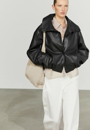 Junge Person trägt eine lockere schwarze Lederjacke, ein beiges Hemd, weiße Hosen und trägt eine große beige Tragetasche auf der Schulter.
