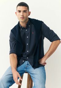 Joven sentado en un taburete de madera, vistiendo una camisa azul marino con las mangas remangadas, un suéter oscuro caído sobre los hombros y jeans de color azul claro.