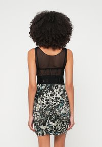 Žena s kovrčavom kosom koja nosi crni mrežasti gornji dio bez rukava i naboranu suknju sa uzorkom, stoji okrenuta leđima prema jednostavnoj pozadini.