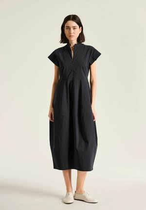 Femme debout portant une robe midi noire à manches courtes avec une fermeture éclair à l'avant et des chaussures beiges à lacets sur un fond uni.