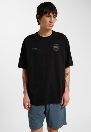 Jonge man met tatoeages staat in een zwart T-shirt met DBU-logo en blauwe Hummel shorts tegen een effen witte achtergrond.