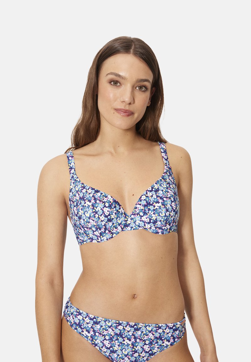 Mujer con un sujetador azul con estampado floral y braguitas a juego, de pie contra un fondo blanco liso, mirando ligeramente hacia un lado.